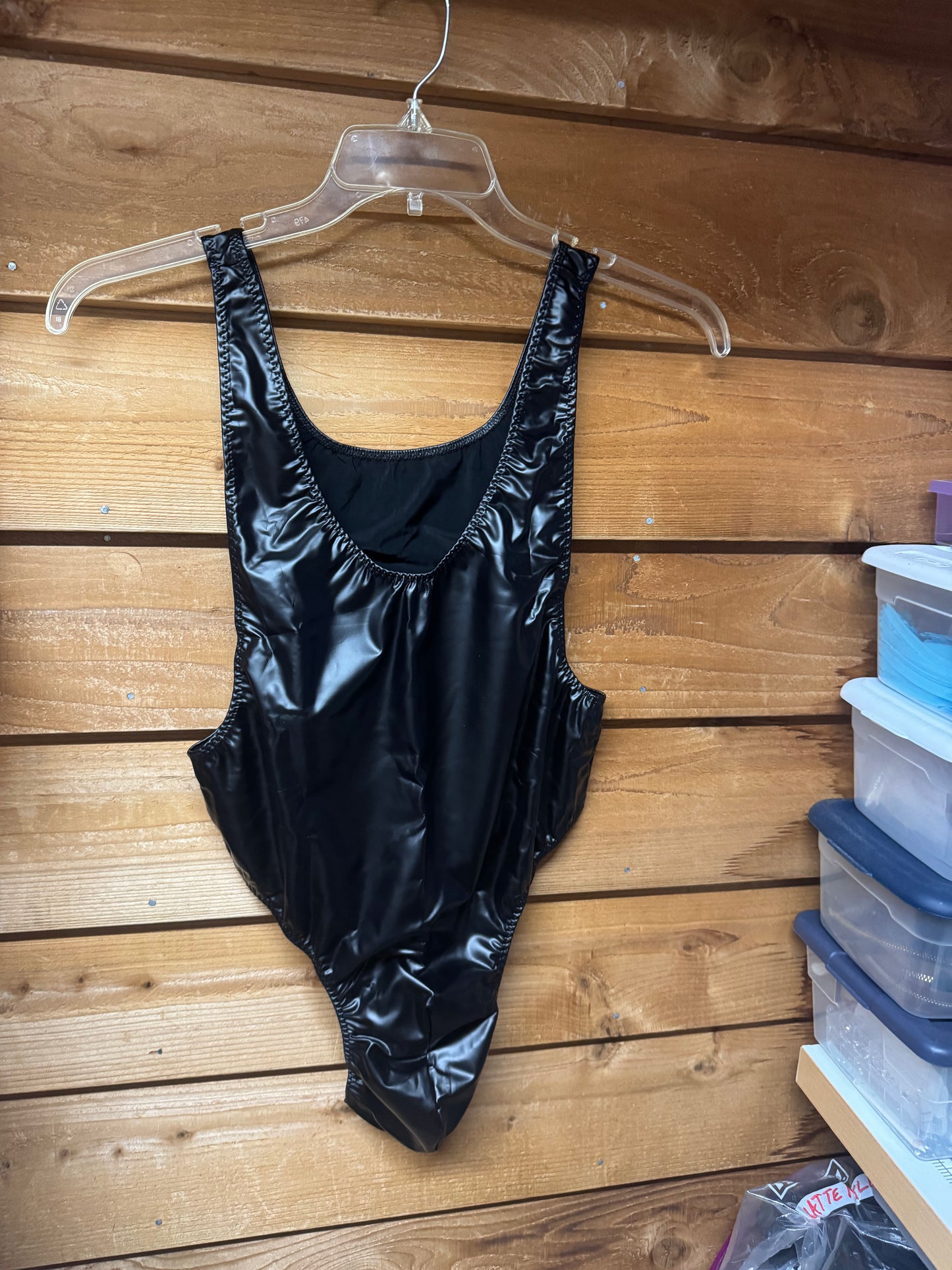 Men's black faux leather thong bodysuit hanging on a clear plastic hanger against a wooden wall.